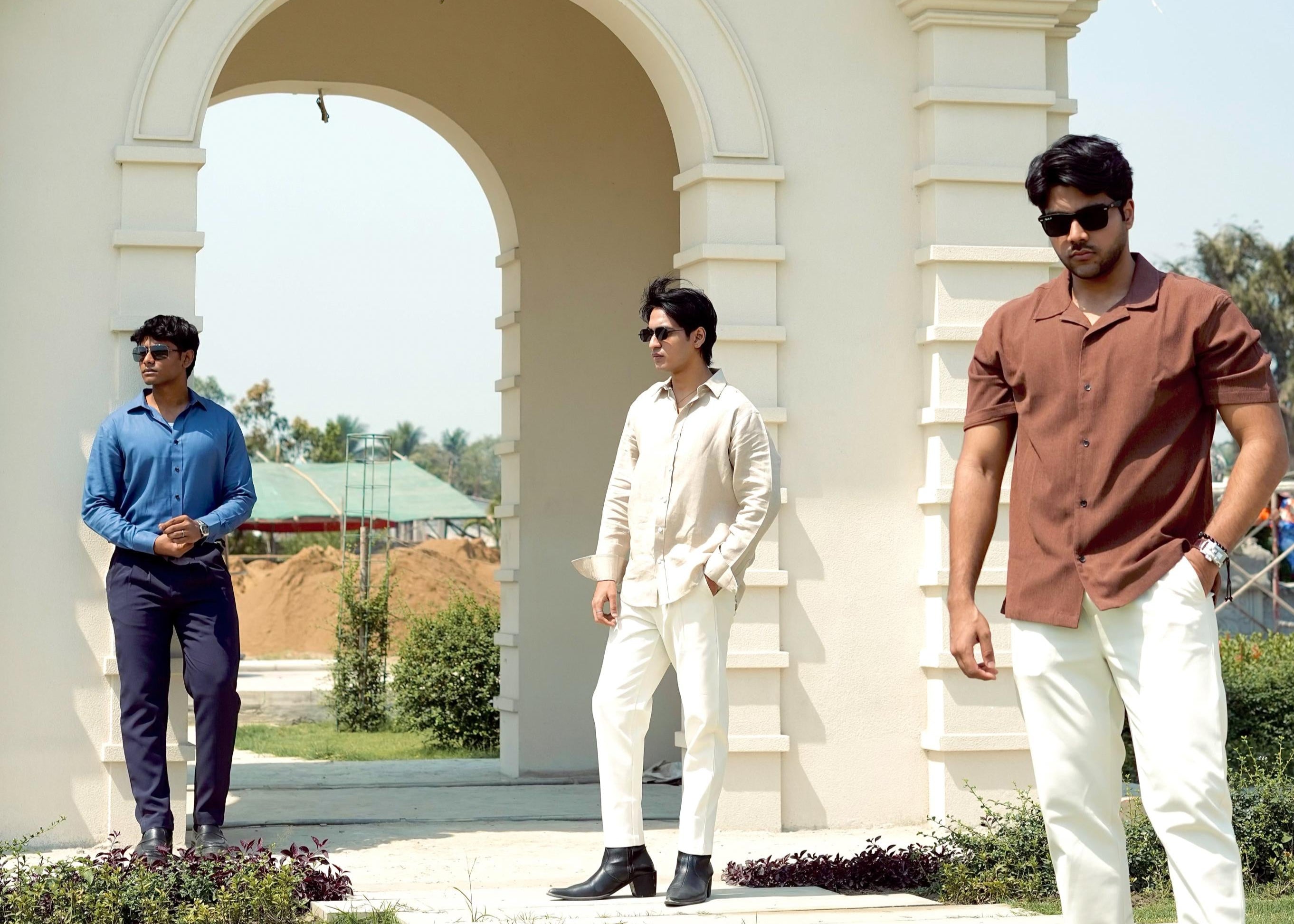 Three men standing in front of a classical architectural archway with greenery in the background.